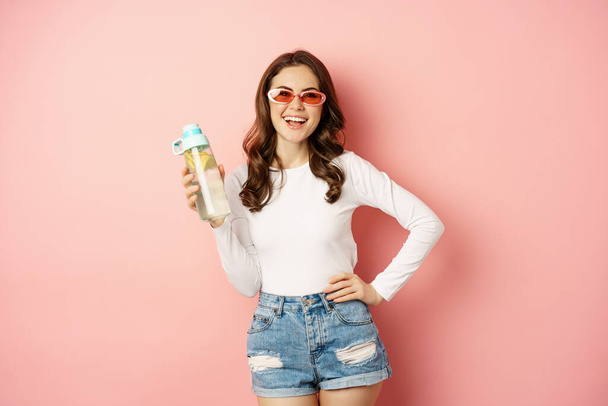Κομψό κορίτσι με ανοιξιάτικο ντύσιμο, φορώντας γυαλιά ηλίου, κρατώντας μπουκάλι νερό με λεμόνι, υγιεινό ποτό, γελώντας και χαμογελώντας, στέκεται πάνω από ροζ φόντο - Φωτογραφία, εικόνα