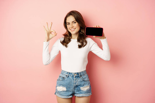 Mujer joven con estilo presentando tienda de la empresa, aplicación móvil, mostrando el teléfono inteligente horizontalmente y bien, buena señal, sonriendo feliz, de pie sobre el fondo rosa - Foto, Imagen