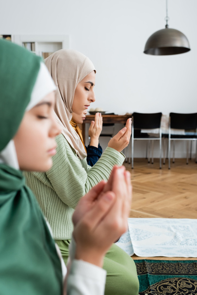 Vista lateral da mulher árabe em roupas tradicionais rezando perto da família em casa  - Foto, Imagem