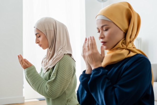 Young arabian woman praying near blurred asian mom at home  - Фото, изображение