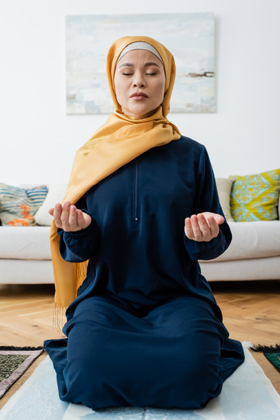 Asian woman praying with closed eyes on traditional rug at home  - Photo, image