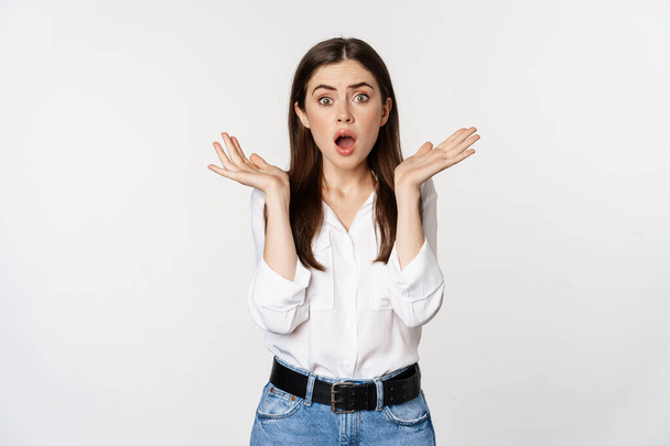 Portrait of concerned female model looking shocked, surprised face reaction, standing in shirt over white background - Foto, immagini