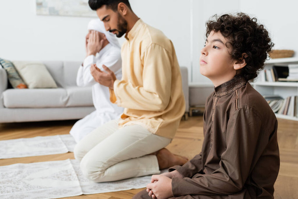 Curly muslim boy praying near blurred father and granddad on rugs at home  - Photo, Image