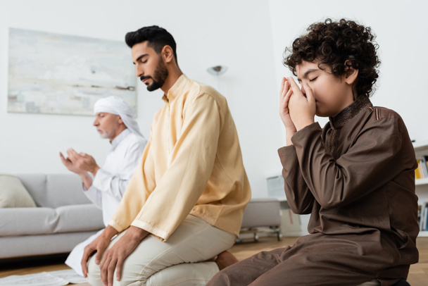 Muslim boy holding hands near face while praying near multiethnic family at home  - Foto, afbeelding