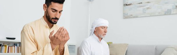 Young arabian man in traditional clothes praying near mature dad at home, banner  - Fotó, kép