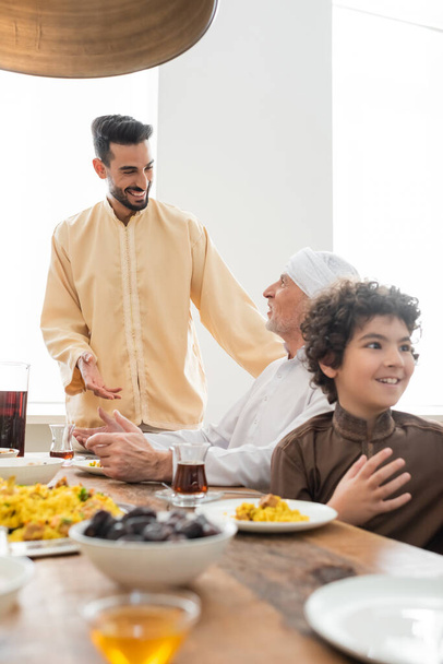 Uśmiechnięty muzułmanin rozmawia z dojrzałym ojcem przy smacznym jedzeniu w domu  - Zdjęcie, obraz