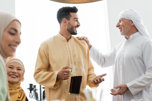 izgatott arab férfi kezében kancsó tea, miközben beszél apa közelében fajok közötti család - Fotó, kép