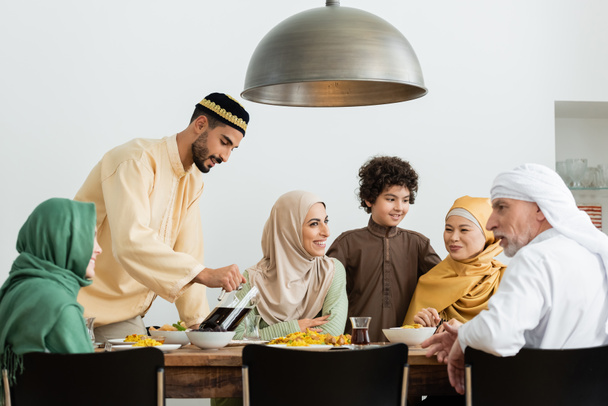 junger arabischer Mann mit Totenkopf gießt Tee während des Abendessens mit interrassischer muslimischer Familie - Foto, Bild