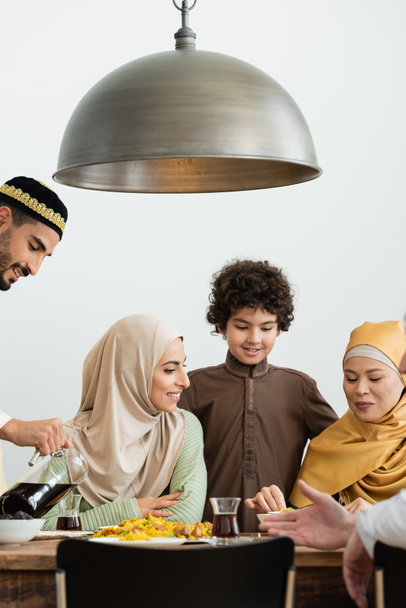 uomo arabo sorridente in skullcap versando tè vicino alla famiglia multietnica - Foto, immagini