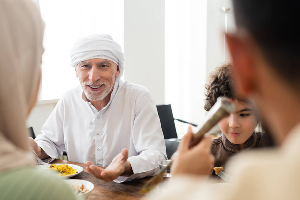 smiling muslim man pointing with hand during dinner with arabian grandson and blurred family - Foto, Imagem