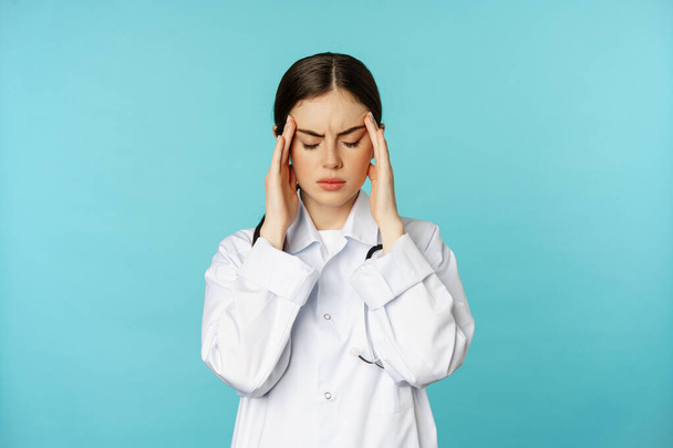 Beeld van een vrouwelijke arts, medisch personeel grimmig van ongemak, hoofdpijn, migraine, gevoel van duizeligheid, staande over torquoise achtergrond - Foto, afbeelding