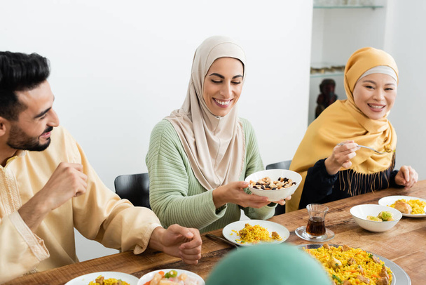 pleased arabian woman in hijab holding bowl with nuts near asian mother and husband - Photo, Image