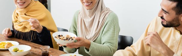 χαμογελαστή γυναίκα κρατώντας μπολ με καρύδια κατά τη διάρκεια του δείπνου με την οικογένεια μουσουλμάνων, banner - Φωτογραφία, εικόνα