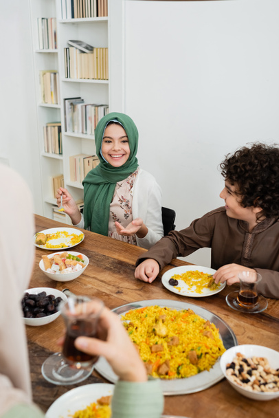 chica feliz en hijab señalando con la mano durante la cena con la familia musulmana - Foto, Imagen