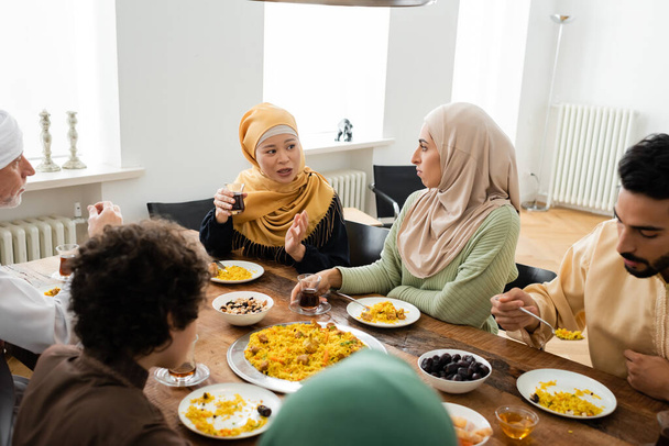 mujer asiática de mediana edad señalando con la mano mientras habla con la familia musulmana multiétnica durante la cena - Foto, Imagen