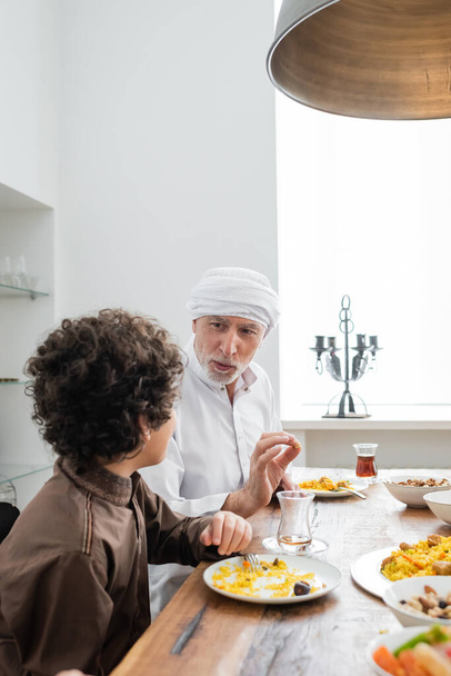 érett muszlim férfi gesztikulál, miközben beszél arab unoka alatt családi vacsora - Fotó, kép