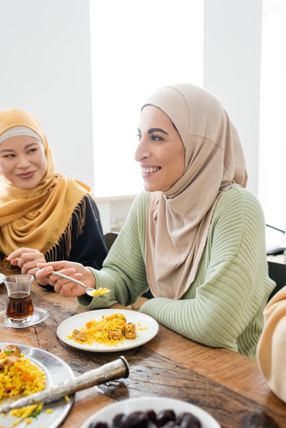 happy muslim woman eating pilaf near asian mother during family dinner - Photo, Image