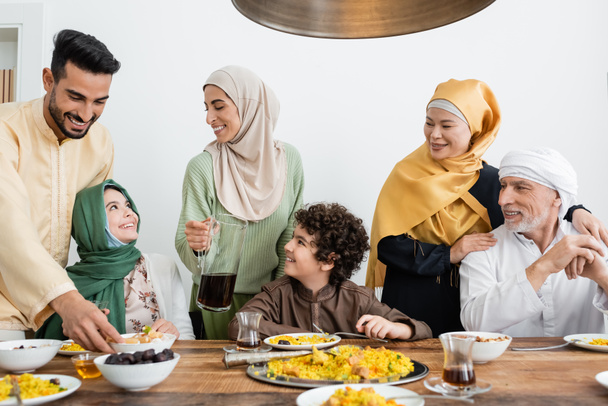 jeune femme arabe tenant une cruche de thé près de la famille musulmane multiethnique en train de dîner à la maison - Photo, image