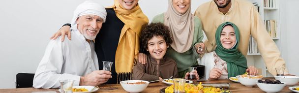 multi-etnische moslim familie glimlachen op camera in de buurt van pilaf en thee op tafel, banner - Foto, afbeelding
