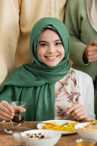happy muslim girl in hijab looking at camera while holding glass of tea near parents - Fotó, kép
