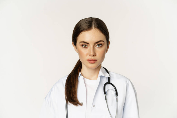 Nahaufnahme Porträt, Gesicht der jungen Ärztin mit Stethoskop, seriös dreinblickend, professionelles medizinisches Personal in der Klinik, vor weißem Hintergrund stehend - Foto, Bild
