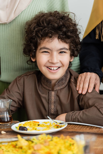 allegro ragazzo arabo musulmano guardando la fotocamera vicino pilaf durante la cena di famiglia - Foto, immagini
