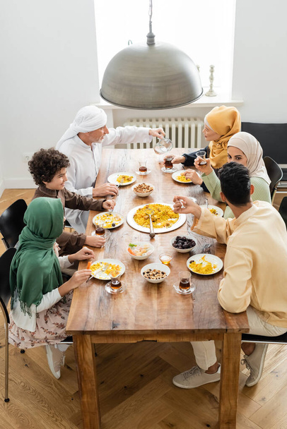 high angle view of middle aged muslim man pouring tea during dinner with multiethnic family - Foto, imagen