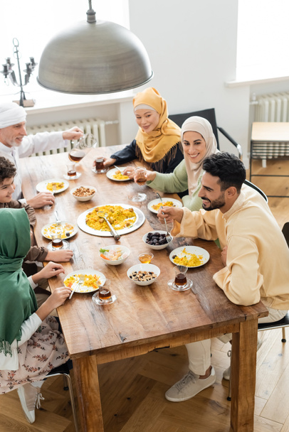 άποψη υψηλής γωνίας ώριμος άνθρωπος ρίχνει τσάι κοντά πολυπολιτισμική μουσουλμανική οικογένεια έχουν δείπνο στο σπίτι - Φωτογραφία, εικόνα
