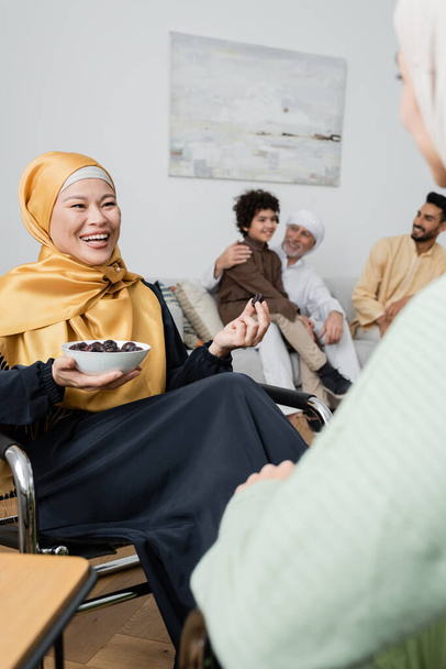 feliz asiático mujer en hijab celebración bowl con fechas cerca borrosa hija y multiétnico musulmán familia - Foto, Imagen