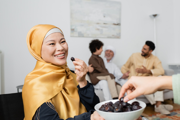 szczęśliwy Azji kobieta w hidżab gospodarstwa daty w pobliżu córka i międzyrasowy muzułmańska rodzina na rozmyte tło - Zdjęcie, obraz