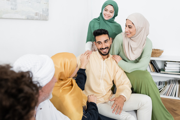 arabski człowiek uśmiechnięty w pobliżu wielokulturowej rodziny muzułmańskiej na kanapie w domu - Zdjęcie, obraz