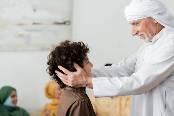 szczęśliwy muzułmanin w turbanowym przytulaniu głowy arabskiego wnuka w pobliżu rozmytej wieloetnicznej rodziny - Zdjęcie, obraz