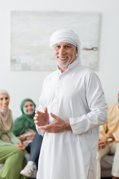 gelukkig moslim man in witte tulband kijken naar camera in de buurt wazig familie - Foto, afbeelding