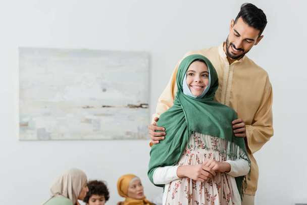 szczęśliwy arabski mężczyzna przytula muzułmańską córkę w pobliżu zamazanej wieloetnicznej rodziny w domu - Zdjęcie, obraz
