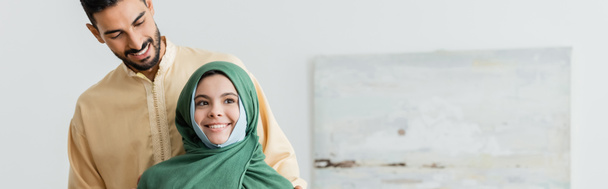 muslim girl in hijab smiling near arabian father at home, banner - Foto, Imagem