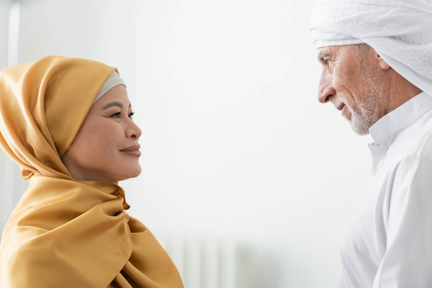 вид сбоку межрасовой мусульманской пары в традиционной одежде, улыбающейся друг другу дома - Фото, изображение