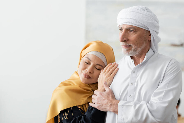 happy middle aged asian woman with closed eyes leaning on muslim husband at home - Фото, изображение