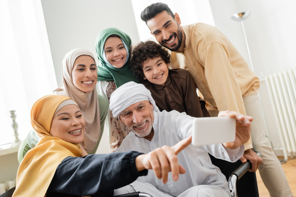 allegra donna asiatica che punta con il dito vicino uomo musulmano prendendo selfie con la famiglia interrazziale - Foto, immagini