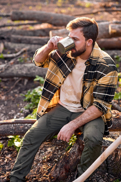 Young guy lumberman sit drinking tea having rest after chopping trees - Zdjęcie, obraz