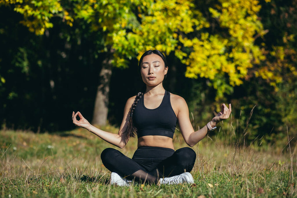 Young beautiful asian woman performs fitness yoga exercises, sitting in lotus position on the grass and meditating in the park - Fotografie, Obrázek