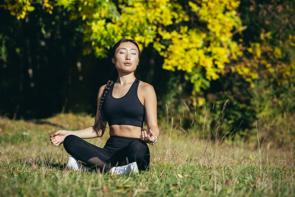 Young beautiful asian woman performs fitness yoga exercises, sitting in lotus position on the grass and meditating in the park - Foto, immagini
