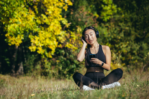 Young beautiful Asian woman sitting in lotus position in the park, relaxing after fitness and running, listening to music on headphones, and using mobile app on phone for fitness - Foto, Imagen