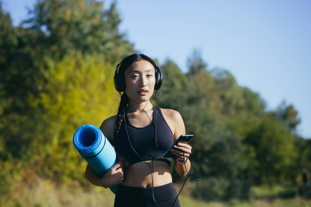 Portret van een mooie slanke Aziatische vrouw, atleet in het bos, luisterend naar muziek met koptelefoon en een yogamat vasthoudend, atleet glimlachend en kijkend naar de camera - Foto, afbeelding