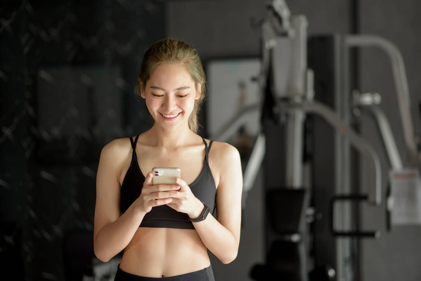 A beautiful smiling woman using a phone while in a gym, Fitness and technology concept . - Фото, изображение
