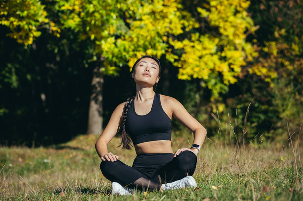 young beautiful sports girl asian woman meditating in park, sitting lotus pose practicing yoga mat, zen. relaxes outdoors in nature in the morning Concept healthy lifestyle, relax, calm, meditation - 写真・画像