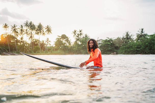 Black long-haired teen man floating on long surfboard, waiting for a wave ready for surfing with palm grove litted sunset rays. Extreme water sports and traveling to exotic countries concept. - Photo, image