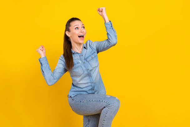 Photo of adorable lucky young lady dressed jeans shirt looking empty space rising fists smiling isolated yellow color background - Foto, immagini