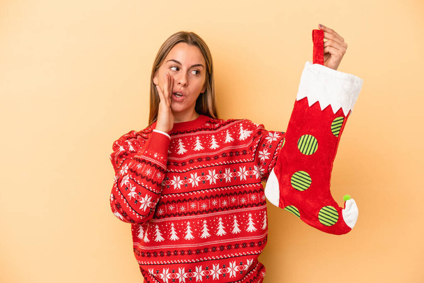 Young caucasian woman holding a elf sock isolated on yellow background is saying a secret hot braking news and looking aside - Photo, Image