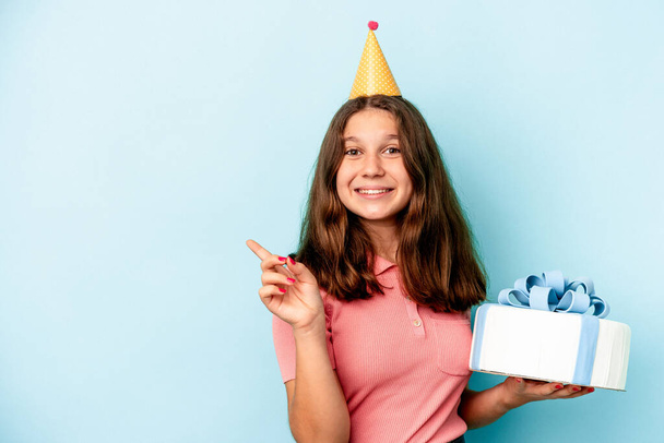 Little caucasian girl celebrating her birthday holding a cake isolated on blue background smiling and pointing aside, showing something at blank space. - Foto, imagen