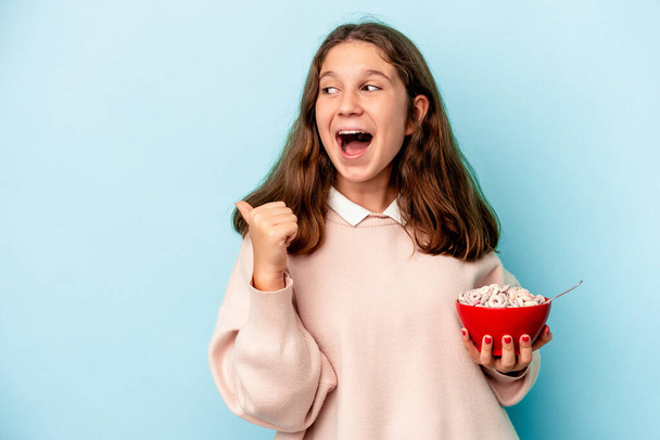 Little caucasian girl holding a bowl of cereals isolated on blue background points with thumb finger away, laughing and carefree. - Photo, Image